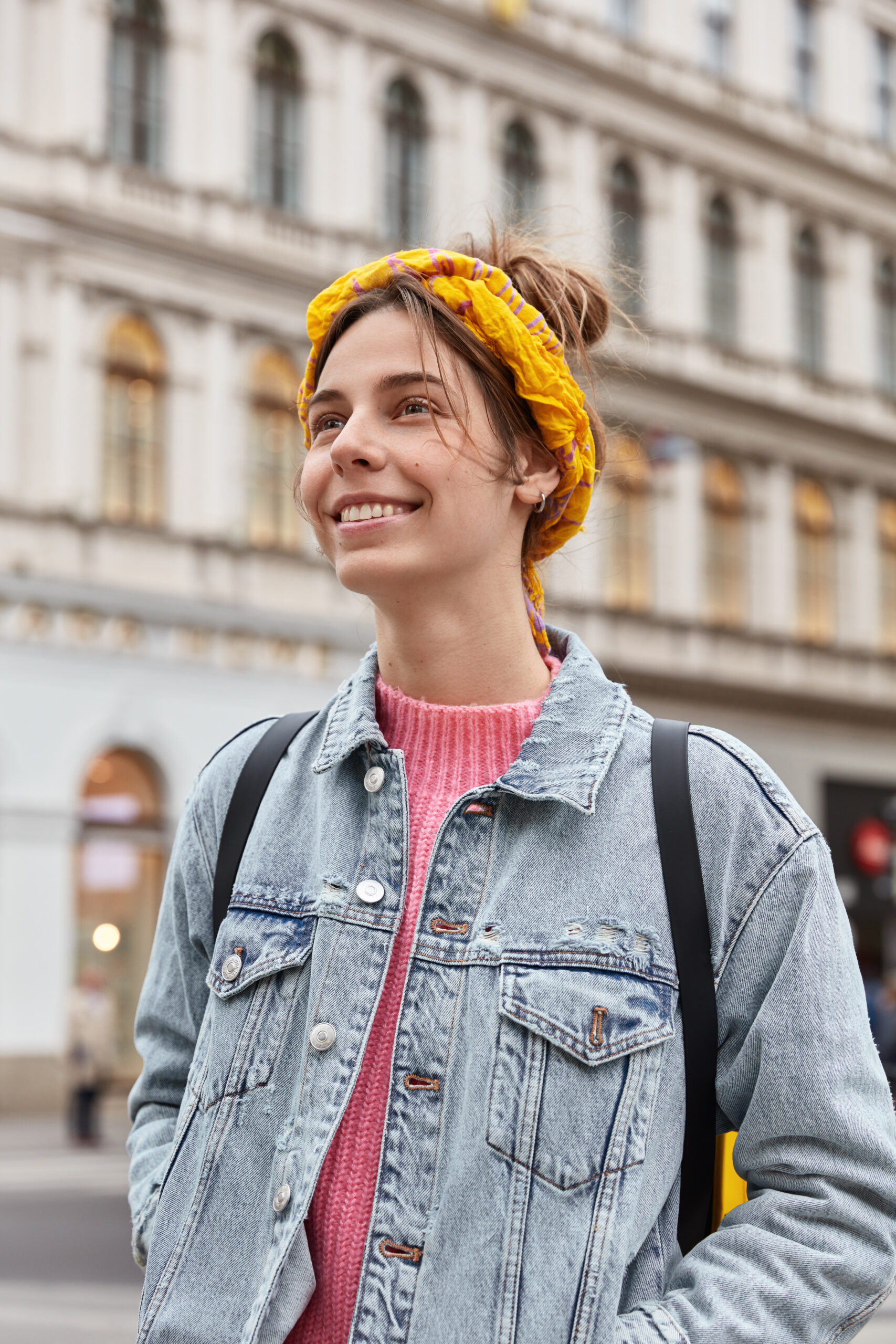 Zdjęcie portretowe uśmiechniętej młodej kobiety, patrzącej w bok na tle klasycznego, zabytkowego budynku w mieście. Kobieta ma na sobie jasną, spraną kurtkę jeansową, różowy sweter oraz jaskrawożółtą, wzorzystą opaskę (turban) na głowie. Na plecach ma czarny plecak. Zdjęcie jest zrobione z dołu, w ciągu dnia.